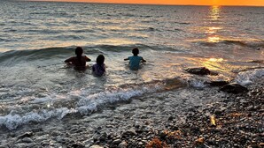 Ein Sonnenuntergang über einem felsigen Strand, drei Personen im Wasser.