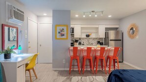 A modern kitchen with white cabinets, a stainless steel refrigerator, and a dining area with orange chairs. - Montrose Amble Studio! Medical Center 02 ★★★★★ (Houston)