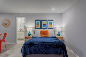 A bedroom with a bed, two nightstands, a red chair, a mirror, and three framed artworks on the wall.