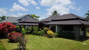 A residential area with two houses featuring dark tiled roofs and stone walls, surrounded by well-maintained lawns and various flowering plants.