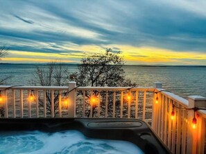 Un jacuzzi con cubierta de madera y luces colgantes, con vista a un cuerpo de agua y un árbol al fondo.