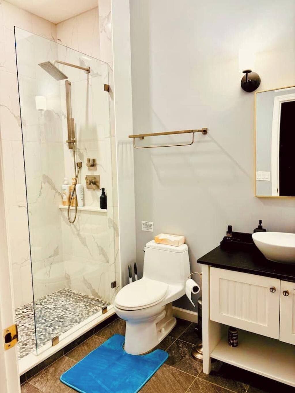Un baño con ducha de mampara de vidrio, inodoro blanco, lavamanos negro y un espejo sobre el lavamanos.