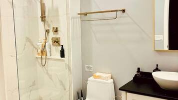 Un baño con ducha de mampara de vidrio, inodoro blanco, lavamanos negro y un espejo sobre el lavamanos.
