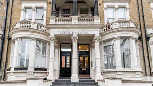 De ingang van Westside Court, een gebouw met een klassieke architectuur, met een opvallende entree met een zwarte deur, witte kolommen en een balkon met sierlijke metalen hekwerk.