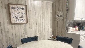 A round dining table with four dark blue chairs, a wooden wall with a 'Welcome to our Emerald Isle beach house' sign, and a kitchen area with a coffee maker and cabinets. - Lucky Enough! Adorable apartment sleeps 5, no cleaning fee/ free beach parking! (Emerald Isle)