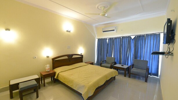 A hotel room with a bed, bedside tables, a chair, a small table, a ceiling fan, and a TV mounted on the wall. - Shikra Tourist Resort (Dabwali)