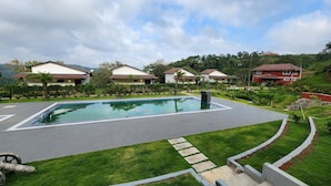 A swimming pool with a surrounding paved area, a grassy lawn, and a row of houses in the background. - Hong Sau Resort (Sakleshpur)