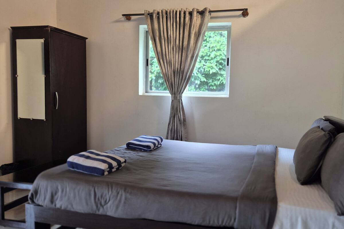 A neatly made bed with pillows and a striped towel, a dark wooden wardrobe, and a window with curtains.