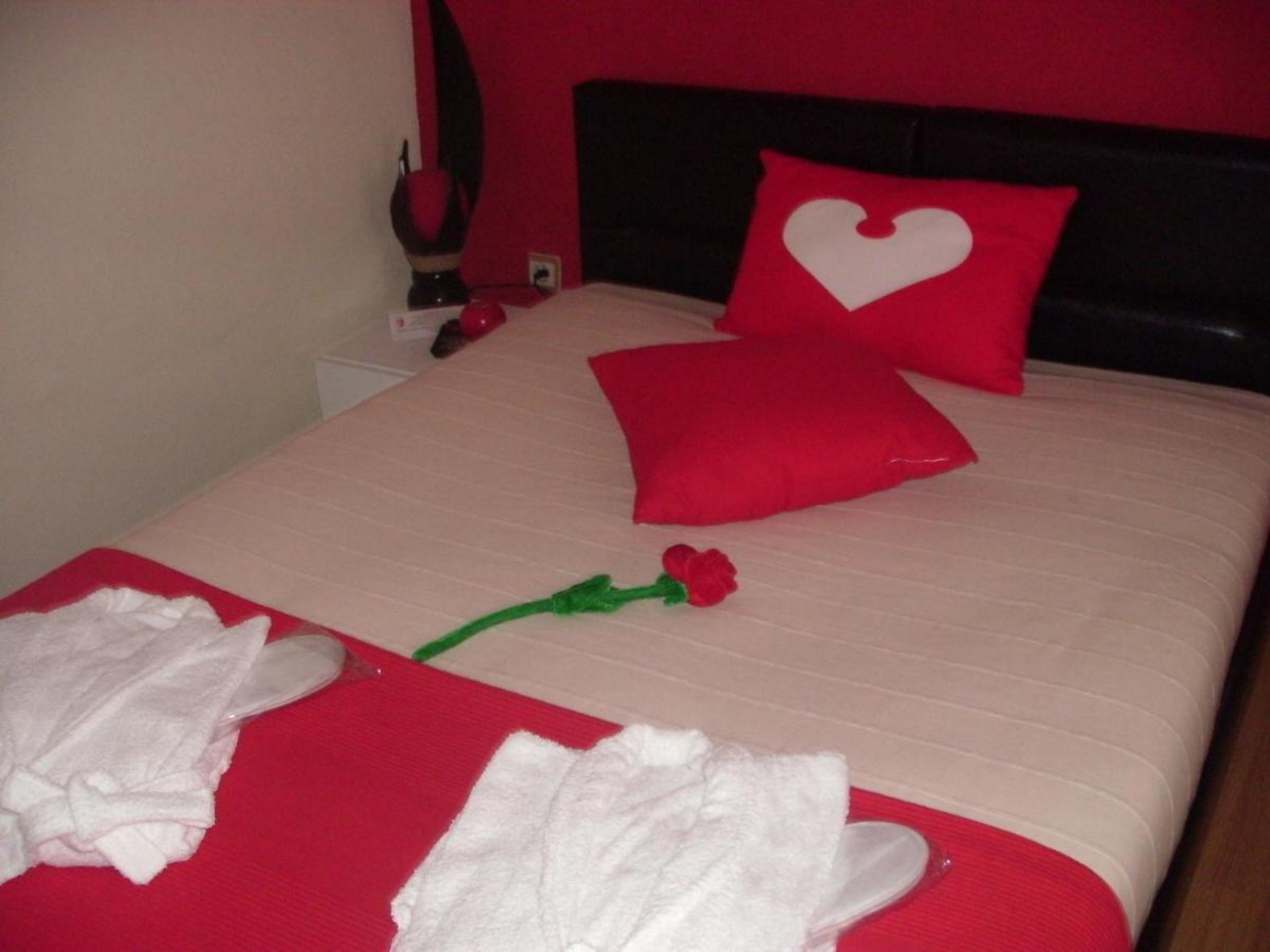 A neatly made bed with red and white bedding, a heart-shaped pillow, and a rose.