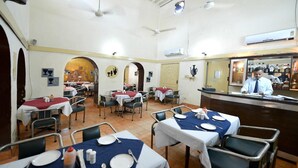 A restaurant with multiple tables, some with red tablecloths and others with white. There are chairs around the tables, and a counter with a person behind it. - Shama Tourist Resort (Gurugram)
