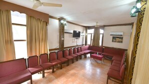 A waiting area with rows of red chairs, a television mounted on the wall, and a ceiling fan. - Shama Tourist Resort (Gurugram)