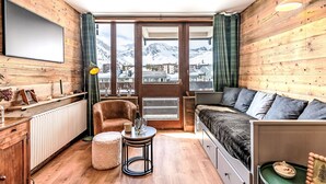 Ein gemütliches Zimmer mit Holzvertäfelung, einer Couch, einem Sessel, einem kleinen Tisch und Blick auf verschneite Berge durch große Fenster.