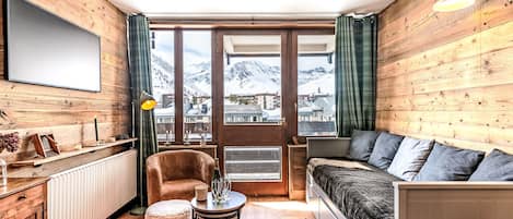 Ein gemütliches Zimmer mit Holzvertäfelung, einer Couch, einem Sessel, einem kleinen Tisch und Blick auf verschneite Berge durch große Fenster.