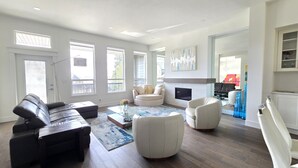 Un vaste salon comprenant un canapé en cuir noir, un fauteuil blanc et un canapé beige. On y trouve une cheminée, un téléviseur fixé au mur et une grande fenêtre qui laisse entrer la lumière naturelle.