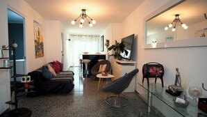 A modern living room with a sofa, armchair, and glass coffee table. There is a TV mounted on the wall, a dining area in the background, and a large mirror above the coffee table.