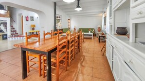 Een eetgedeelte met een lange houten tafel en stoelen, een keuken met witte kastjes, en een trap die naar een hoger gelegen verdieping leidt.