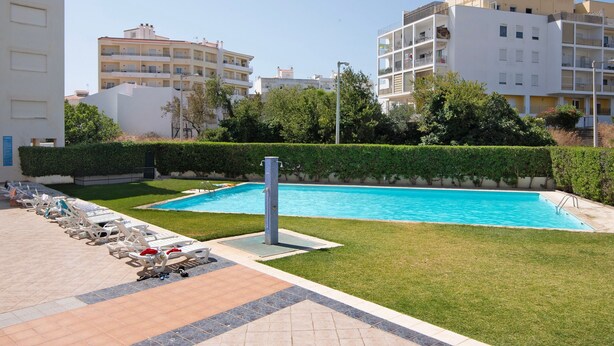 Une piscine avec des chaises longues, un petit espace aménagé en jardin et, au fond, des immeubles à plusieurs étages.