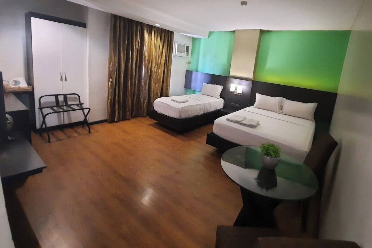 Ein Hotelzimmer mit zwei Betten, einem Schreibtisch, einem Stuhl, einem kleinen Tisch mit Pflanze und einem Vorhang.