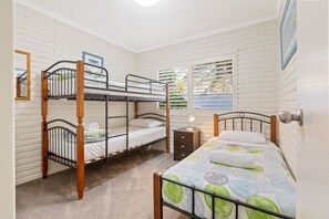 A room with two bunk beds, a single bed, a nightstand, a window with blinds, and a door with a handle. - Beachfront Apartments: Ground floor Fishpen (Merimbula)