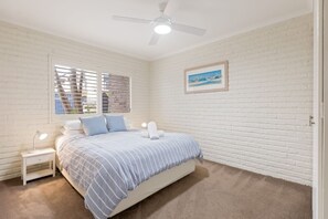 A bedroom with a bed, a nightstand, a ceiling fan, a window with blinds, and a framed picture on the wall. - Beachfront Apartments: Ground floor Fishpen (Merimbula)