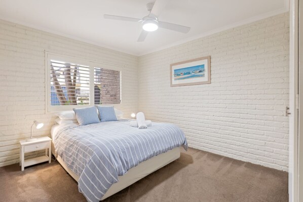 A bedroom with a bed, a nightstand, a ceiling fan, a window with blinds, and a framed picture on the wall. - Beachfront Apartments: Ground floor Fishpen (Merimbula)