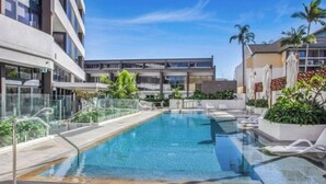 A swimming pool with clear blue water, surrounded by lounge chairs and palm trees. - Modern 1-Bedroom Apartment in Central Gold Coast (Surfers Paradise)