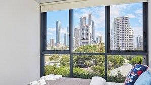 A hotel room with a large window offering a city view, a bed with white linens, and a blue pillow. - Modern 1-Bedroom Apartment in Central Gold Coast (Surfers Paradise)