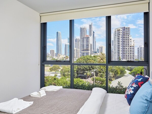 A hotel room with a large window offering a city view, a bed with white linens, and a blue pillow.