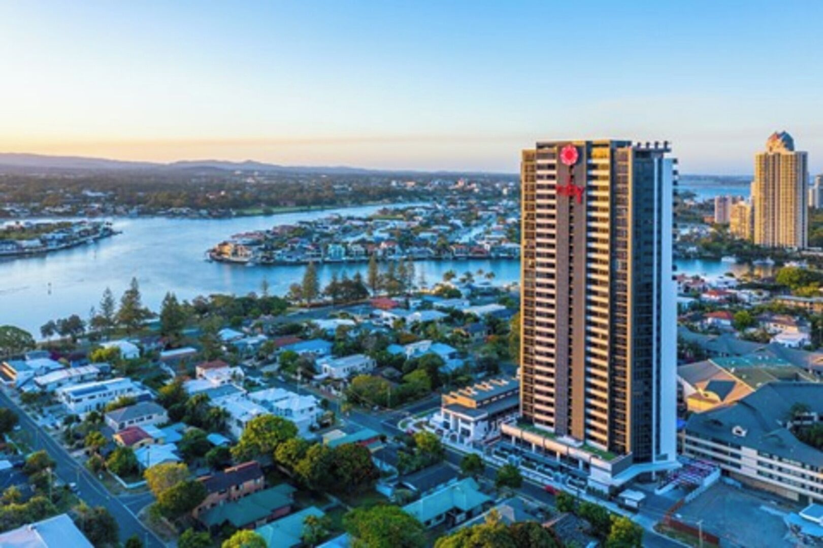 Un hotel alto con un logotipo rojo llamativo, rodeado de una zona residencial y una masa de agua.