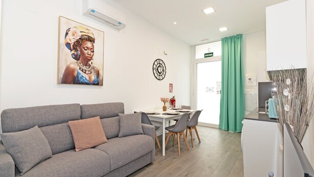 A modern living room with a grey sofa, a dining table with chairs, a clock on the wall, and a kitchen area with a fridge and a sink. - GuestReady - Flat near the Marina of Valencia (Valencia)