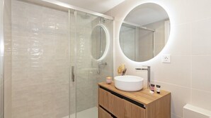 A modern bathroom with a glass-enclosed shower, a round mirror, a wooden vanity, and a white sink. - GuestReady - Flat near the Marina of Valencia (Valencia)
