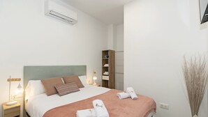 A neatly made bed with a pink blanket, white pillows, and a green headboard. There is a bedside table with a lamp, a wooden shelf with folded towels, and a tall vase with reeds. - GuestReady Essential - Flat near Valencia Marina (Valencia)