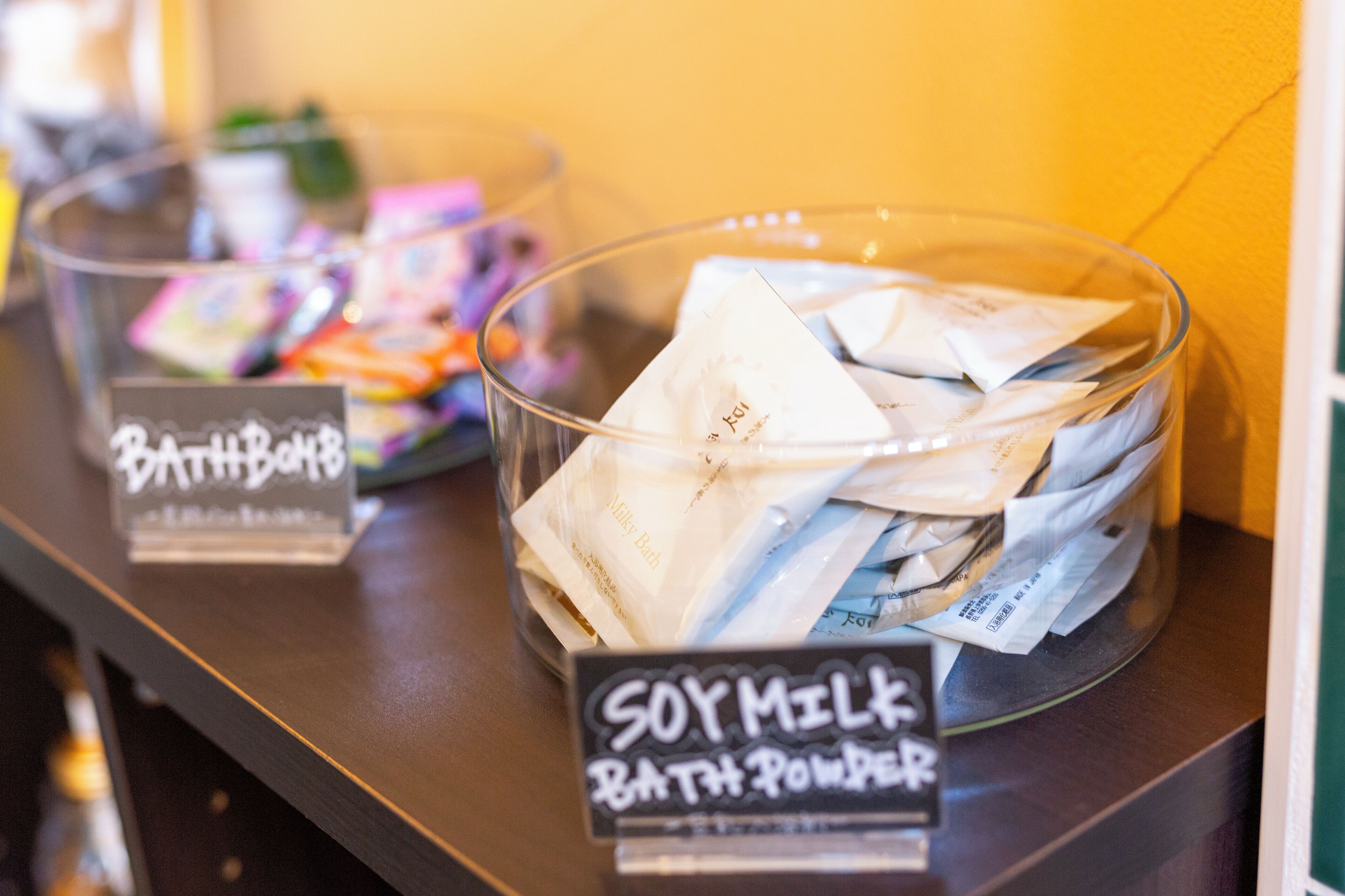 A glass container labeled "SOY MILK BATH POWDER" on a counter with a sign "Bath & Body" in the background.