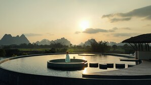 A circular infinity pool with a central fountain, surrounded by a tiled deck, set against a backdrop of mountains and a sunset.