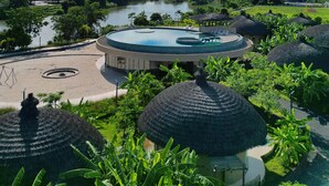 A resort with a circular pool, thatched-roof huts, and a view of a lake and mountains.