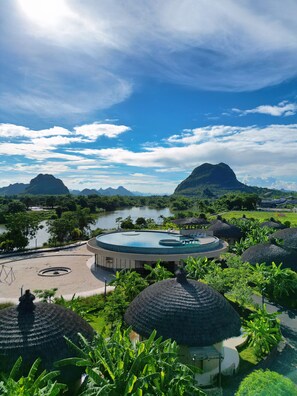 Un resort con piscina circular, cabañas de techo de paja y vistas a un lago y montañas.