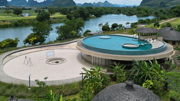A circular swimming pool with a surrounding deck, surrounded by lush greenery and thatched-roof structures.