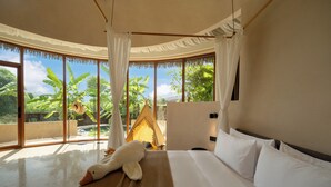A spacious bedroom with a large bed, a view of lush greenery, and a thatched-roof structure outside.
