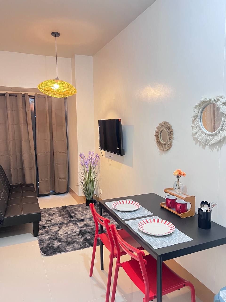 A dining area with a black table, red chairs, a yellow hanging light, and a TV mounted on the wall.