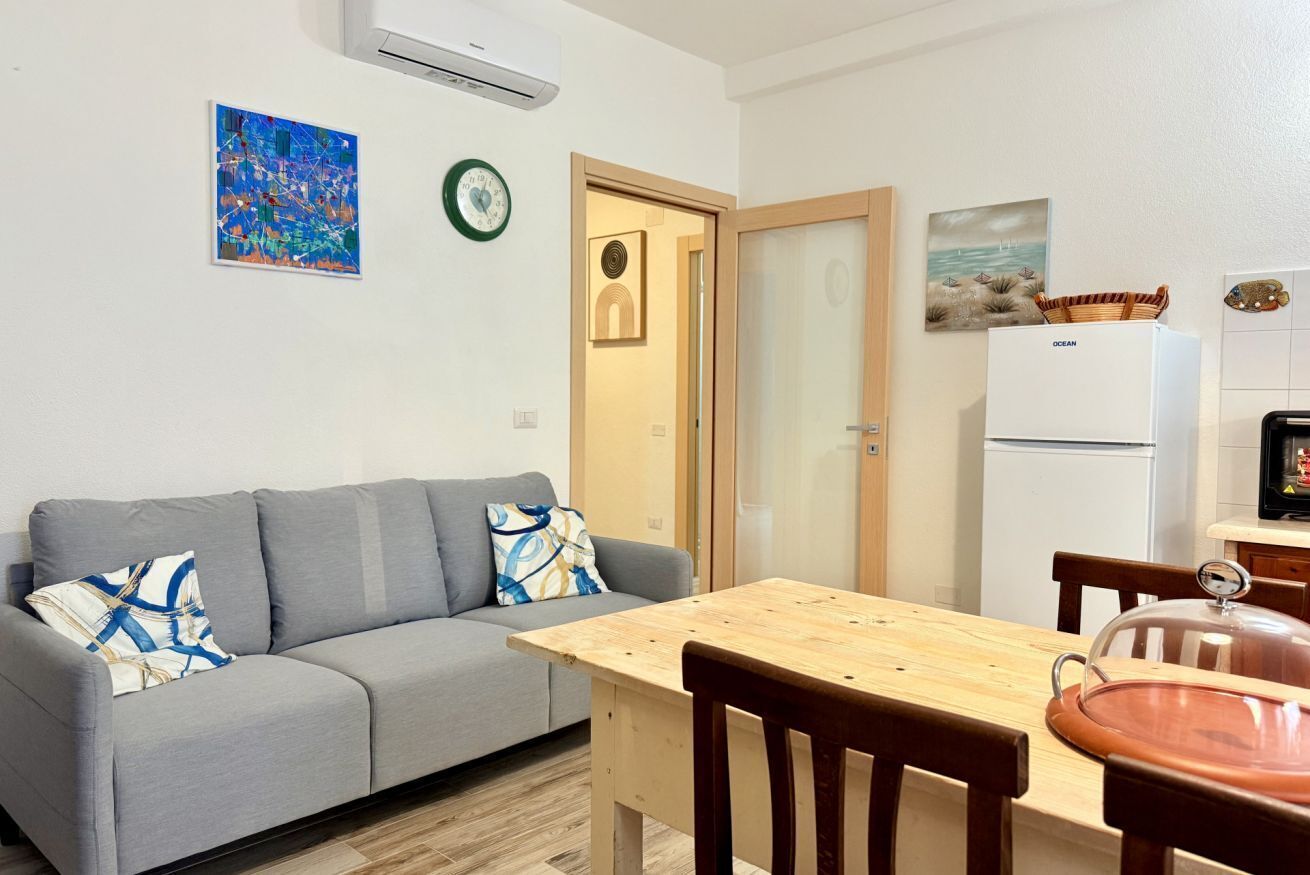 Een moderne woonkamer met een grijze bank, een houten eettafel en een keuken met een koelkast en een fornuis.