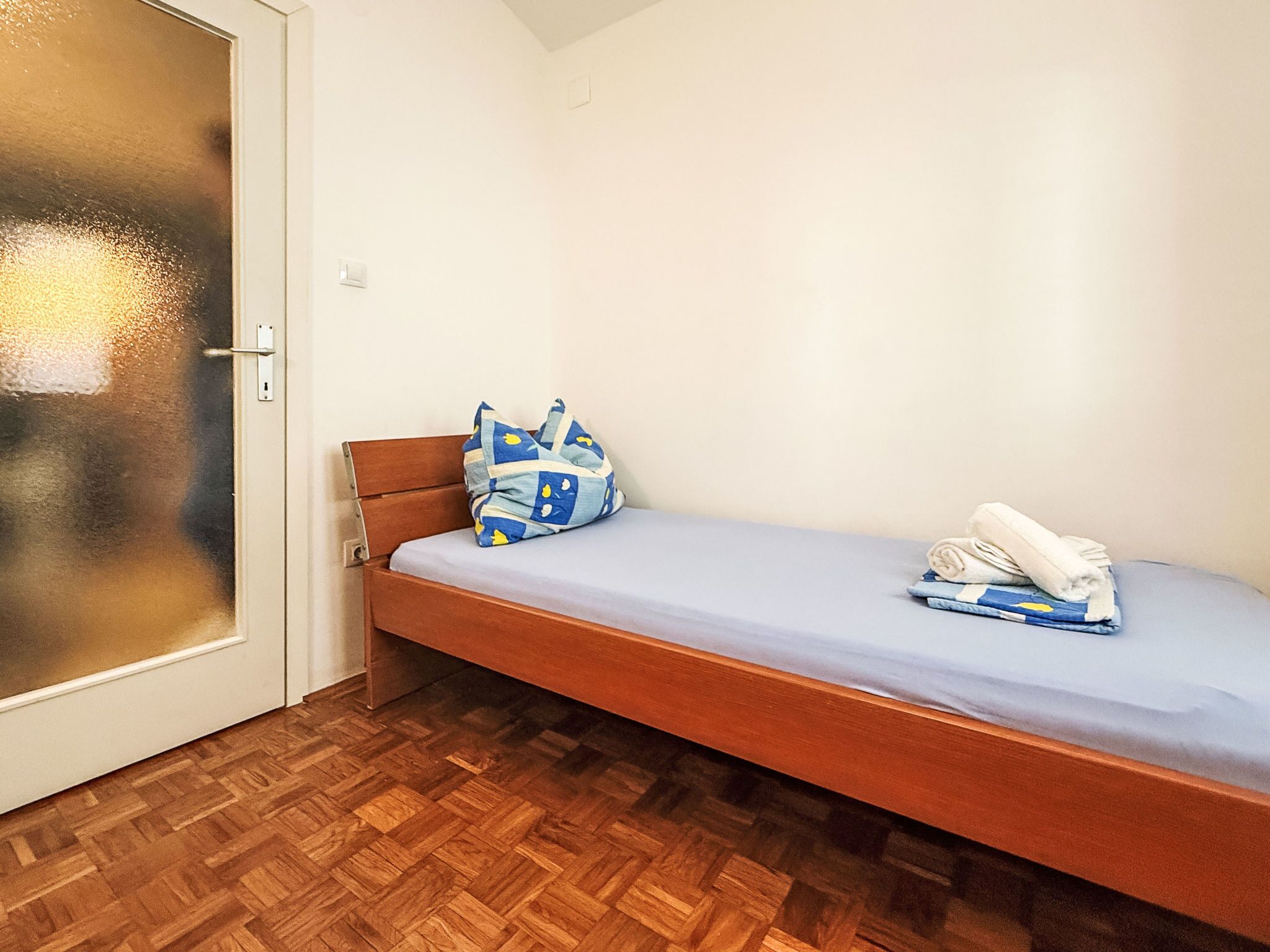 A small, single-bed room with a wooden headboard, a blue patterned pillow, and a folded towel on the bed.