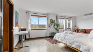 Ein Schlafzimmer mit einem Bett, einem Schreibtisch, einer Zimmerpflanze und großen Fenstern.