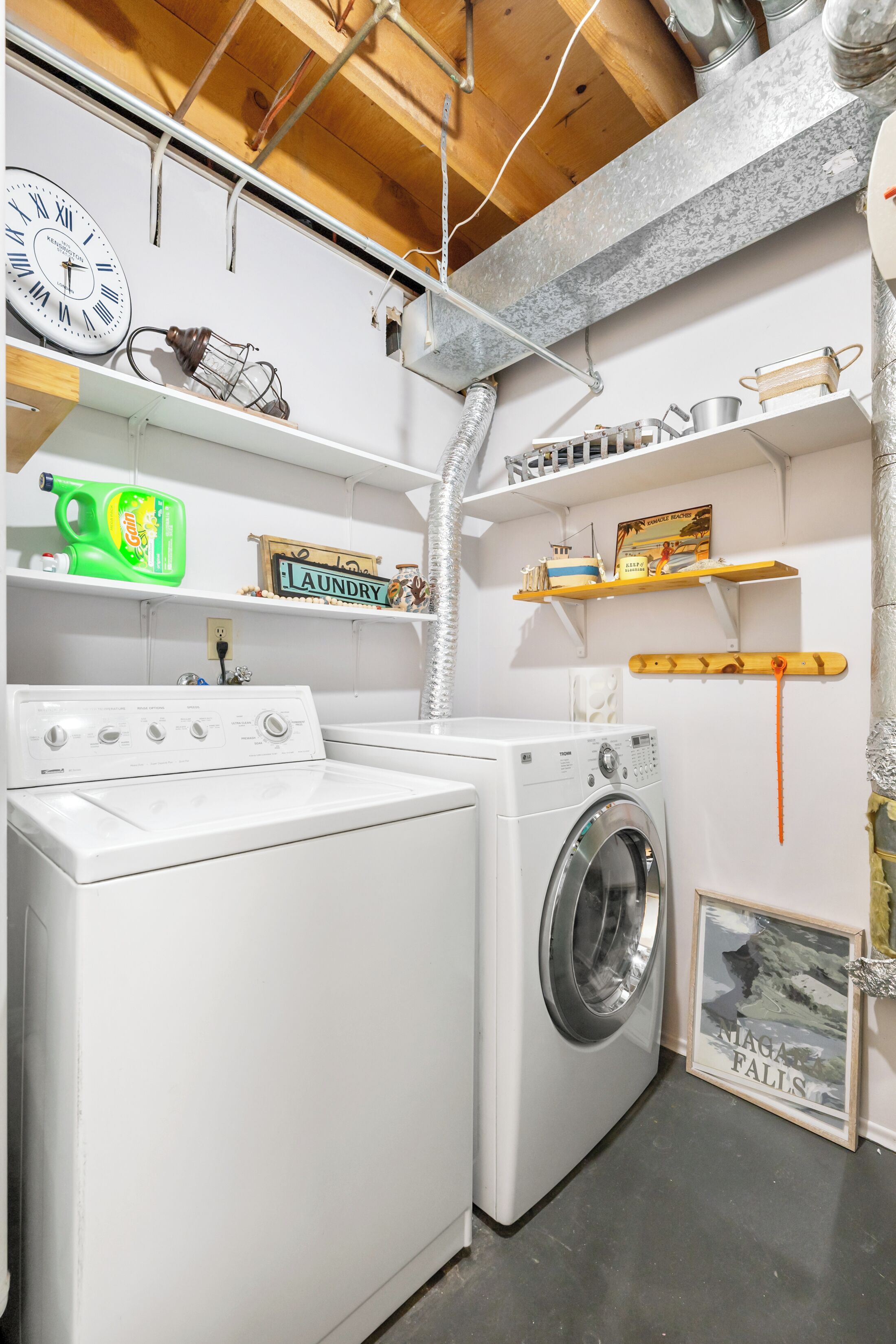 Une buanderie équipée d’un lave-linge et d’un sèche-linge, avec des étagères remplies de divers objets et une horloge au mur.