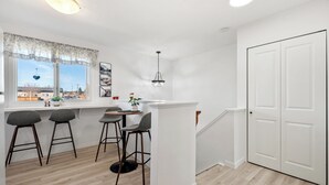 A kitchen with a window, three bar stools, a hanging light fixture, and a white door. - Charming 2-bedroom condo central NW (Calgary)