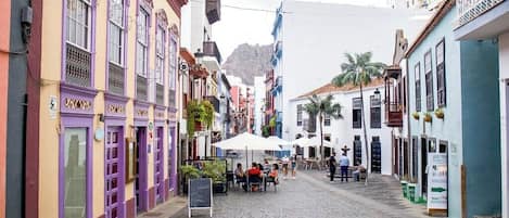 A cobblestone street lined with colorful buildings, outdoor seating areas, and a mountain in the background.
