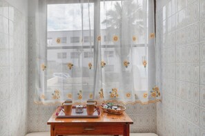 A corner room with patterned tiles, a window with a floral curtain, a wooden table with a tray, and a decorative basket.