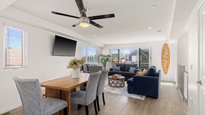 A modern living room with a wooden dining table, grey upholstered chairs, a blue sofa, and a ceiling fan. - Single Family Retreat | Steps to Bay & Beach | Fireworks Views (San Diego)