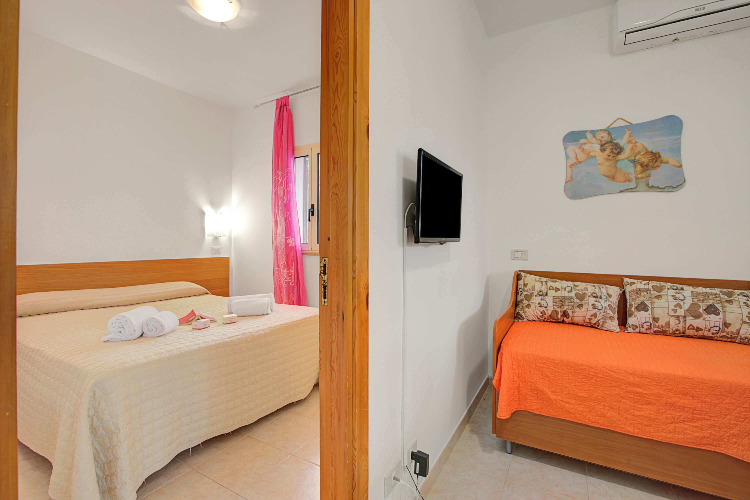 A room with two beds, one with a beige bedspread and the other with an orange bedspread. There is a television mounted on the wall and a painting above it.