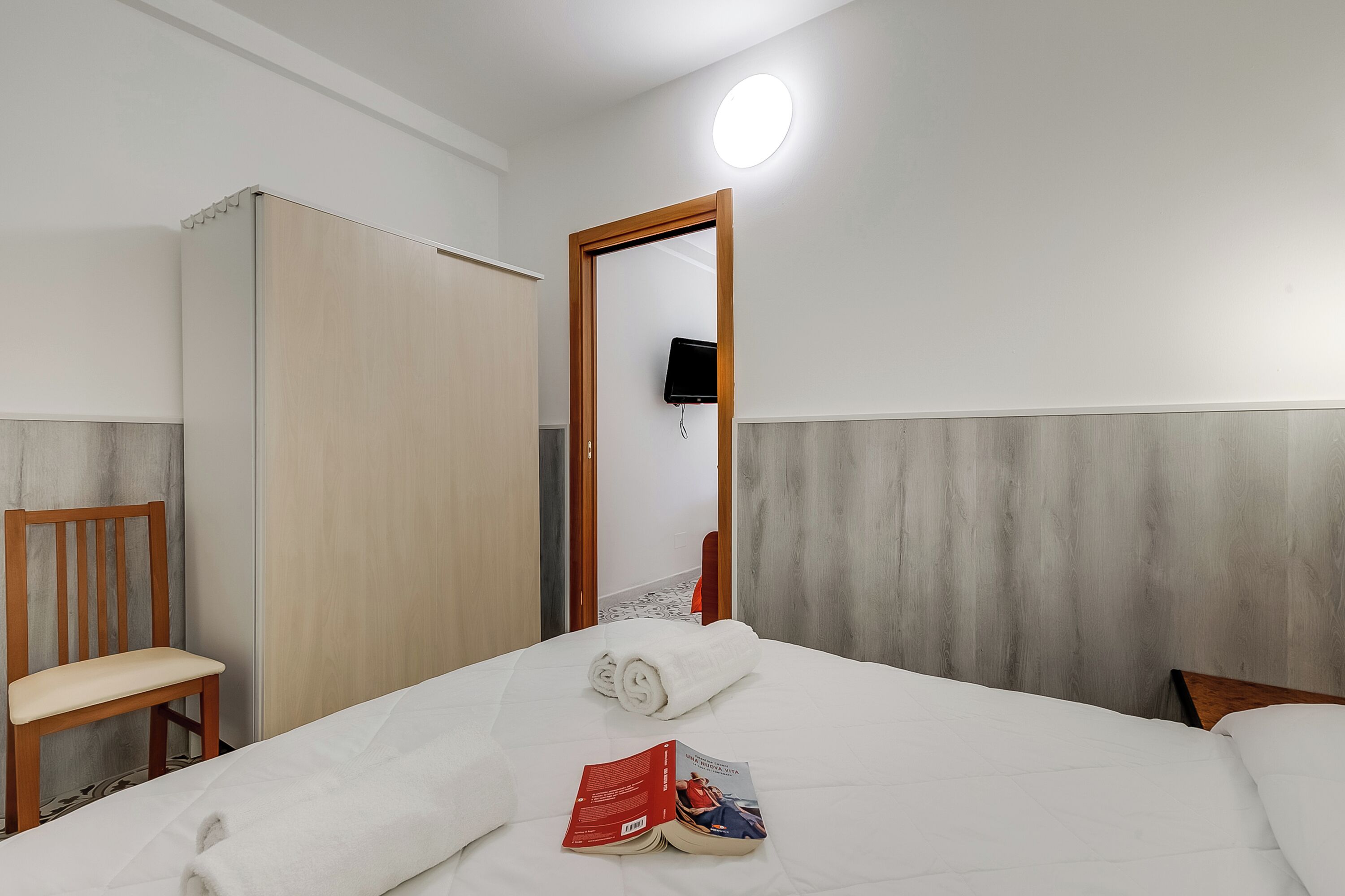 Een hotelkamer met een bed, een houten stoel, een kledingkast en een flatscreen televisie aan de muur.