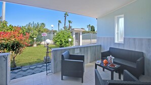 Ein Balkon mit Korbmöbeln, einem kleinen Tisch mit Getränken und Obst, und Blick auf einen Garten und ein Gebäude im Hintergrund.